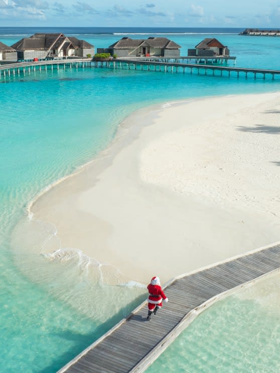 Santa Claus walking along a wooden bridge over turquoise waters towards a white sand beach at Niyama Private Islands Maldives