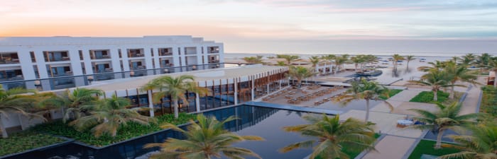 Resort building surrounded by palm trees and water features with view of the beach at sunrise
